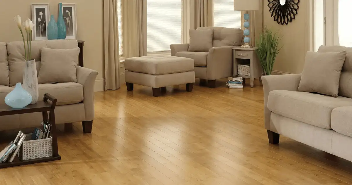 A living room with professionally installed bamboo flooring, beige sofas, a matching ottoman, a side table with a blue lamp, vases, framed pictures, and large windows with blinds and curtains in Long Beach, CA