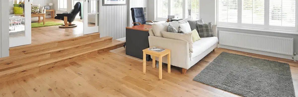 A modern living room with professionally installed laminate flooring, a beige sofa, a small side table, a gray rug, and large windows with white shutters in Long Beach, CA